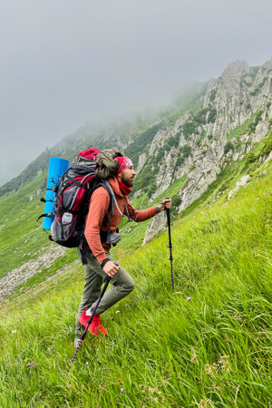 Man hiker climbs steep, grassy slope with trekking poles. Tourist carry large backpack, with blue sleeping pad. Rugged mountain landscape with misty cliffs in background, creating adventurous scene.の写真素材