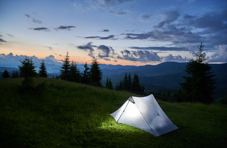 Touristic tent glows warmly on grassy hillside at dusk, surrounded by dark silhouettes of mountains and pine trees. Sky is deep blue with scattered clouds, as last light of sunset fades into night.の写真素材