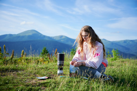 Woman prepares meal from packaged pouch next to portable camping stove. Female tourist with long...