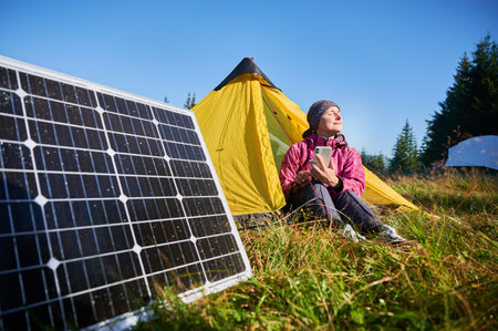 Happy woman sits in front of yellow tent, charging her phone by using solar panel, highlighting use of renewable energy and eco-friendly camping. On background lush green trees and clear blue sky.の写真素材