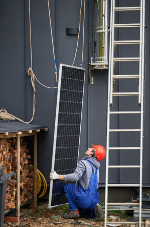 Men installers installing solar panel system on roof of house. Workers in helmets lifting up photovoltaic solar module with help of ropes outdoors. Concept of alternative and renewable energy.の写真素材