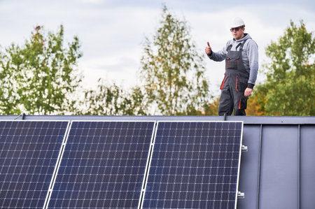 Portrait of man technician installing photovoltaic solar modules on roof of house, showing thumbs up. Electrician building solar panel system outdoors. Concept of alternative and renewable energy.の写真素材