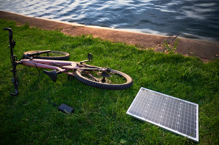 Pink electric mountain bike lies on grass near lakeside, connected to solar panel for charging. Serene scene features bike and solar panel with calm water in background.の写真素材