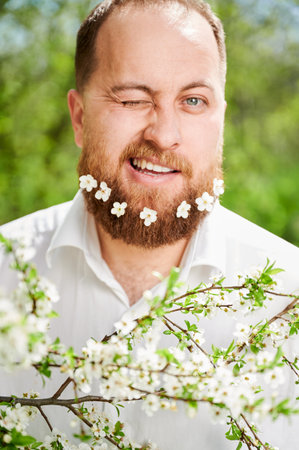 Portrait of happy man with small white flowers dotted across his beard, winking one eye in front of blossom tree at springtime.の写真素材
