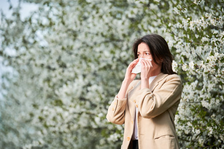 Woman allergic suffering from seasonal allergy at spring in blossoming garden at springtime. Young woman sneezing and blowing nose with nasal handkerchief in front of blooming tree. Allergy concept.の写真素材