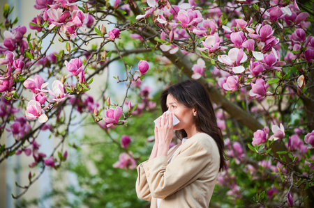 Woman allergic suffering from seasonal allergy at spring in blossoming garden at springtime. Young woman sneezing and blowing nose with nasal handkerchief in front of blooming tree. Allergy concept.の写真素材