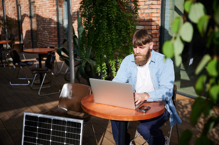 Man works on laptop and smartphone at outdoor table. Solar panel nearby, emphasizing sustainable, modern eco-friendly workspace that combines technology and sustainability.の写真素材