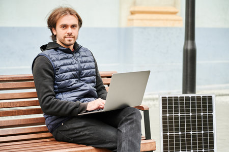 Man sits on wooden bench, working on laptop connected to solar panel. Urban setting and casual attire highlight modern, sustainable, mobile, eco-friendly workspace.の写真素材