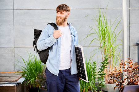 Bearded man carrying solar panel and black backpack, stands in front of a modern building, smiling. Happy guy in light blue shirt and white t-shirt surrounded by greenery and plants.の写真素材