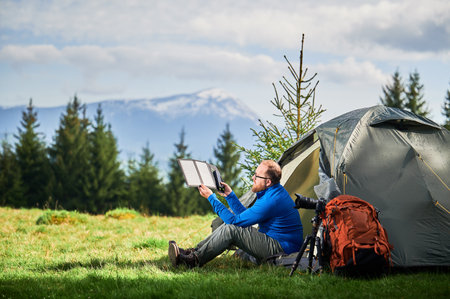 Man sits beside tourist tent, holding portable solar panel charger to capture sunlight for charging power bank. Large orange backpack, camera on tripod. Snow-capped mountains under partly cloudy sky.の写真素材