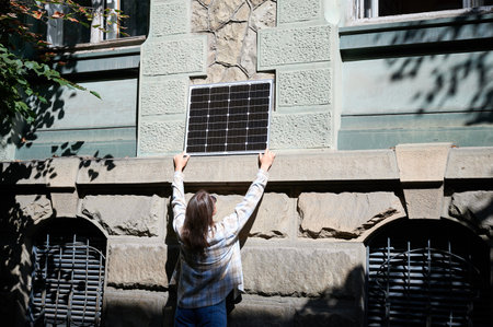 Woman holding photovoltaic solar panel in front of historical building, wearing plaid shirt and sunglasses. Concept of integration of sustainable renewable energy sources into old architecture.の写真素材