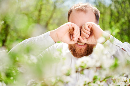 Man allergic suffering from seasonal allergy at spring in blossoming garden at springtime. Close up of bearded man sneezing and feeling itchy eyes in front of blooming tree. Spring allergy concept.の写真素材