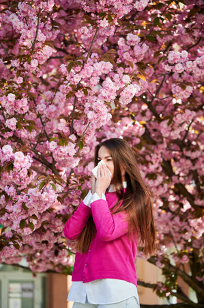Woman allergic suffering from seasonal allergy at spring in blossoming garden at springtime. Woman sneezing and blowing nose using nasal handkerchief in front of blooming tree. Spring allergy concept.の写真素材