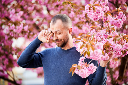 Man allergic suffering from seasonal allergy at spring in blossoming garden at springtime. Bearded man feeling itchy eyes in front of blooming tree. Spring allergy concept.の写真素材