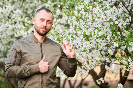 Man allergic using medical nasal drops, suffering from seasonal allergy at spring in blossoming garden. Handsome smiling man showing thumbs up near blooming tree outdoors. Spring allergy concept.の写真素材