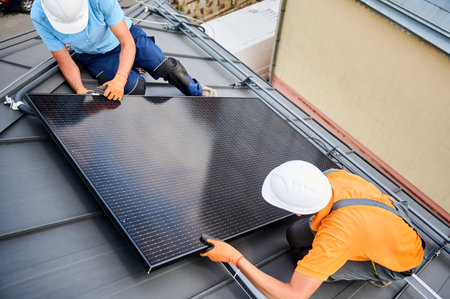 Workers building photovoltaic solar panel system on rooftop of house. Men technicians in helmets and gloves installing solar module with help of hex key outdoors. Renewable energy generation concept.の写真素材