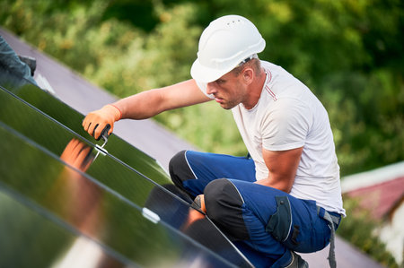 Worker building photovoltaic solar panel system on rooftop of house. Man engineer in helmets and gloves installing solar module with help of hex key outdoors. Alternative energy generation concept.の写真素材