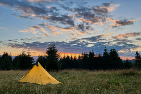 Yellow tourist tent set up in grassy field at sunset, surrounded by trees in evening. Sky painted with vibrant hues of orange, pink, and purple as sun sets, casting warm glow over serene campsite.の写真素材