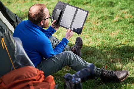 Man sits beside tourist tent, holding portable solar panel charger to capture sunlight for charging power bank. Renewable energy in outdoor activities, blending technology with natural environment.の写真素材