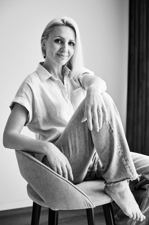Pretty female relaxing on stool. Portrait of woman with blonde hair smiling straight to camera. Beautiful woman sitting on stool in her apartment. Black and white image.の写真素材