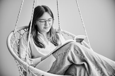 Beautiful girl relaxing and reading book on indoor swing. Pretty female in glasses excited about story she reading. Cozy afternoon with book in hanging chair in home. Black and white image.の写真素材
