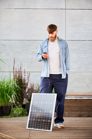 Bearded man using smartphone connected to photovoltaic solar panel. Integration of sustainable renewable energy into everyday life, demonstrating practical use of solar power for charging devices.の写真素材