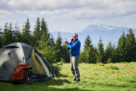 Tourist stands beside tent, talking on phone and holding portable solar panel. Orange backpack and camera tripod nearby. Snow-capped mountains and lush evergreen trees under partly cloudy sky.の写真素材