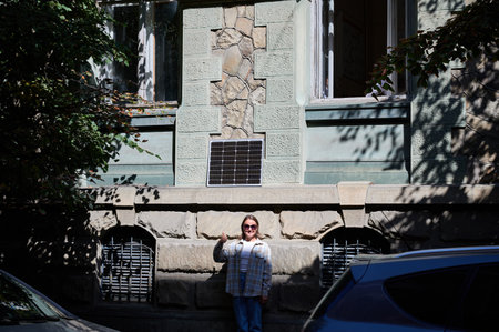 Happy woman gives thumbs-up while standing in front of building with solar panel mounted on wall. She wears sunglasses, white top, plaid shirt. Scene partially shaded, highlighting urban environment.の写真素材