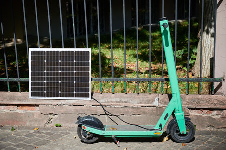Mint-green electric scooter parked on stone pavement next to solar panel leaning against iron fence. Solar panel connected to scooter, emphasizing sustainable method of charging in urban setting.の写真素材