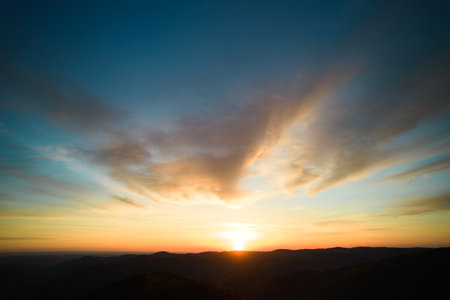 Sun sets behind rolling hills, casting warm, golden glow across sky. Wispy clouds, tinged with hues of orange and yellow, stretch across deepening blue sky. Serene and expansive landscape.の写真素材