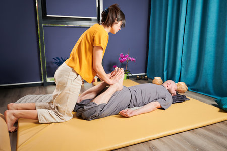 Masseuse making thai yoga massage. Woman therapist doing traditional massage treatment, stretching male patients body. Bald client lying on mat, enjoying hand massage in calm atmosphere.の写真素材