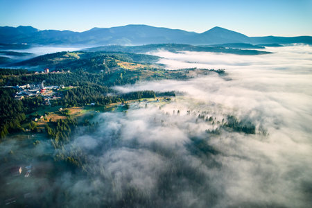 Aerial view of lush, green valley with scattered houses bathed in early morning sunlight. Mist rolls through forested hills, creating ethereal atmosphere against backdrop of distant mountains.の写真素材