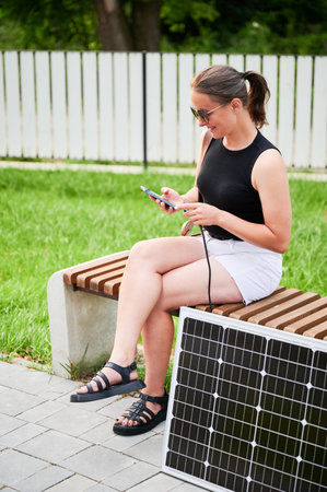 Happy woman using smartphone connected to photovoltaic solar panel. Integration of sustainable renewable energy into everyday life, demonstrating practical use of solar power for charging devices.の写真素材