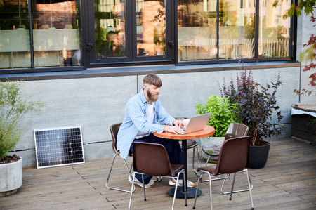 Bearded man works on laptop at outdoor round table, surrounded by greenery. Solar panel nearby, emphasizing sustainable, modern eco-friendly workspace that combines technology and sustainabilityの写真素材