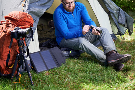 Bearded man sits at entrance of tourist tent in grassy area, charging power bank with portable solar panel. Nearby, tripod-mounted camera and orange backpack indicate his interest in photography.の写真素材