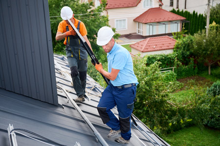 Workers building solar panel system on roof of house. Two men installers in helmets carrying photovoltaic solar module outdoors. Alternative, green and renewable energy generation concept.の写真素材