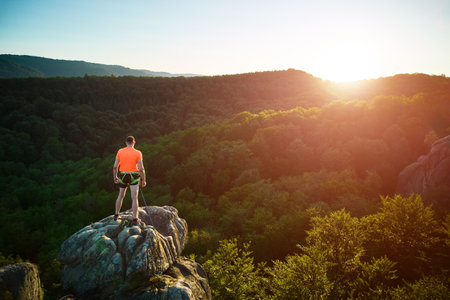 Aerial view of climber standing atop rock formation at Dovbush Rocks in Carpathian mountains, Ukraine. Sun sets, casting warm glow over lush, green forest, distant hills, creating breathtaking view.の写真素材