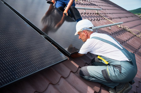 Workers building solar panel system on rooftop of house. Two men installers in helmets installing photovoltaic solar module outdoors. Alternative, green and renewable energy generation concept.の写真素材