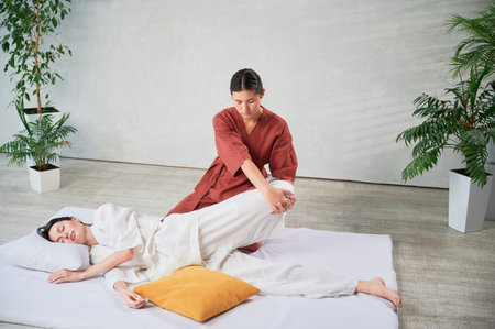 Masseuse making thai yoga massage. Woman therapist doing traditional massage treatment, stretching female patients body. Client lying on mat, enjoying hand massage in calm atmosphere.の写真素材