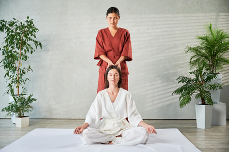 Masseuse making thai yoga massage. Woman therapist doing traditional massage treatment for female patient indoors. Client sitting on mat, meditating, enjoying hand massage in calm atmosphere.の写真素材