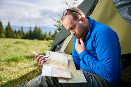Man sits in tourist tents entrance, deeply focused on map. Tourist holds compass and contemplates his route, surrounded by lush green field with trees and mountains in distance under cloudy sky.の写真素材