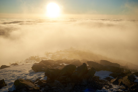 Sunrise over snow-covered mountain peak. Warm golden light of rising sun bathes clouds in soft hues of orange and pink. Snow and scattered rocks in foreground add texture and depth to scene.の写真素材