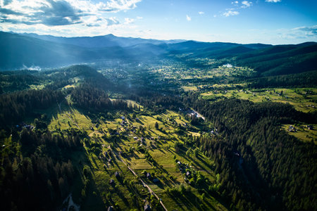 Aerial view of lush, green valley dotted with small houses and winding roads, surrounded by dense forests and rolling hills. Sunlight filters through scattered clouds. Mountains rise under blue sky.の写真素材