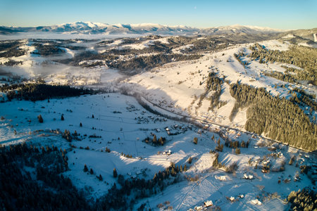 Aerial view of snow-covered valley with winding road passing through small village. Landscape features gently rolling hills, dense forests, and distant mountains under clear blue sky, with soft mist.の写真素材