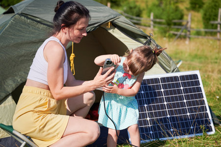 Young child and mother play with smartphone while charging with photovoltaic solar panel near tourist tent in summer. Integration of renewable energy in outdoor camping activities.の写真素材