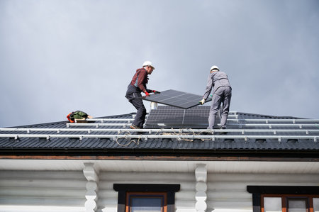 Builders installing solar panel system on roof of house. Men workers in helmets carrying photovoltaic solar module outdoors. Concept of alternative and renewable energy.の写真素材