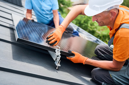 Workers building photovoltaic solar panel system on rooftop of house. Close up of men technicians in helmets and gloves installing solar module with help of hex key outdoors. Renewable energy.の写真素材