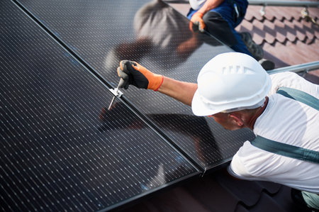 Workers building photovoltaic solar panel system on rooftop of house. Close up of men technicians in helmets and gloves installing solar module with help of hex key outdoors. Alternative energy.の写真素材