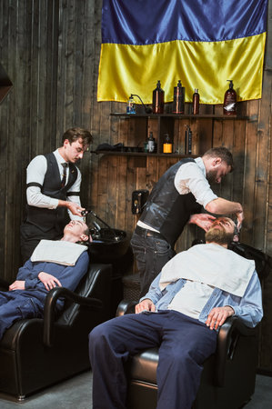 Professional barbers washing hair with deep cleaning shampoo. Busy day in modern Ukrainian barbershop. Male clients relaxing while getting their hair done.の写真素材
