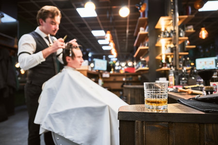 Close-up photo of a glass of whiskey. High-quality barbershop offering drinks to customers. Glass with alcohol on the background of hairstylist making haircut.の写真素材